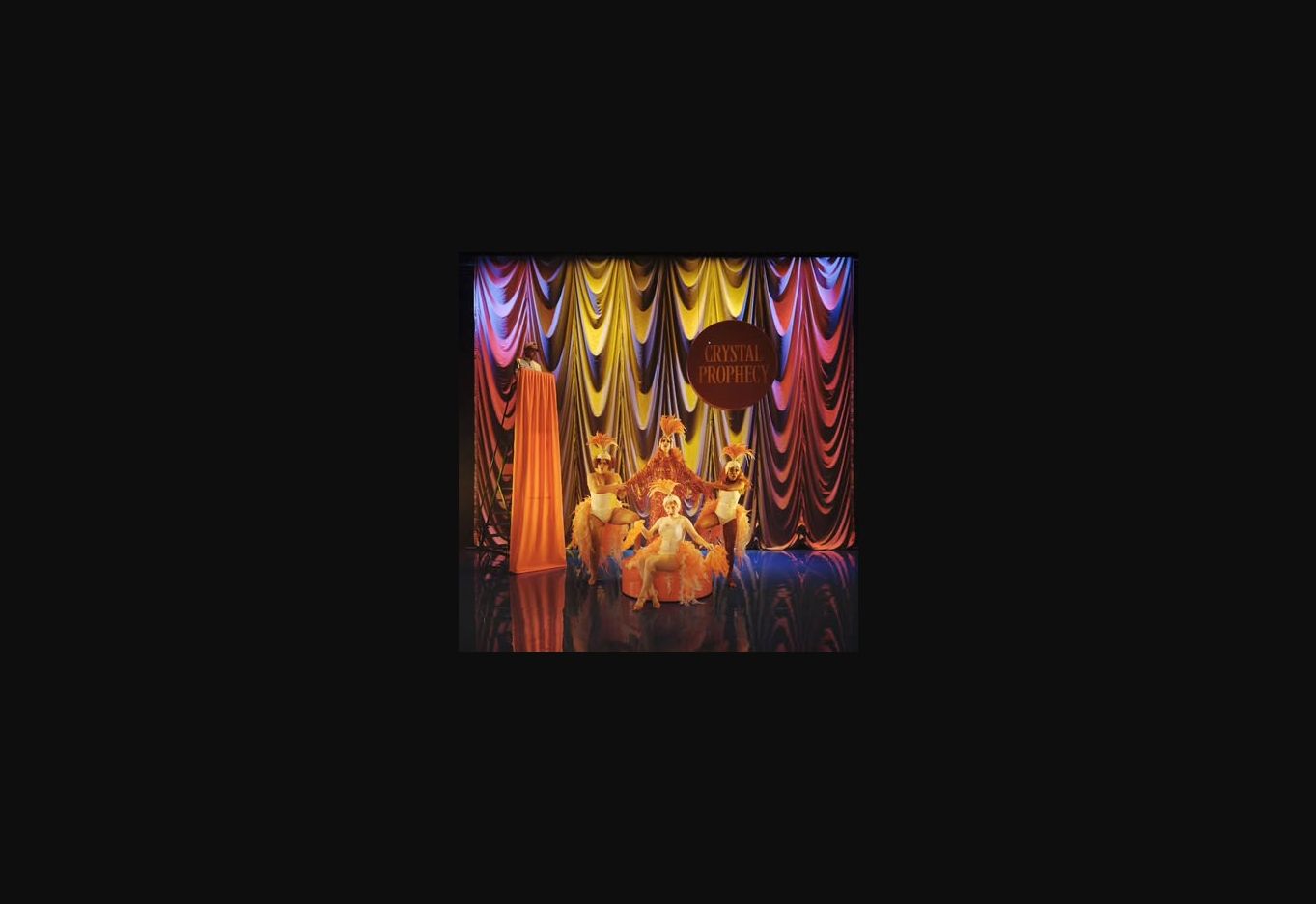 A vividly costumed ensemble performing beneath colourful theatre drapes