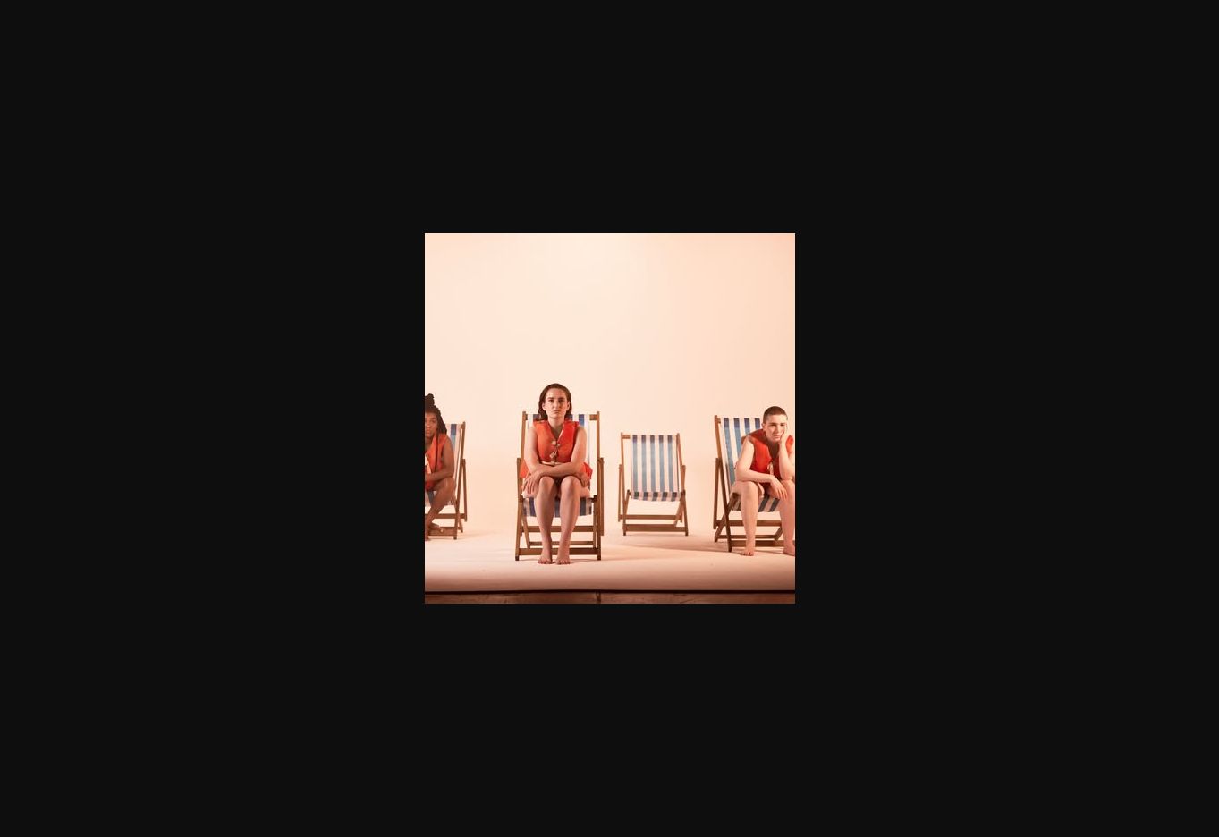 Performers in red costumes seated on deckchairs against a pale stage